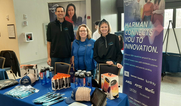 Three people standing behind a promotional booth for Harman at a corporate event, displaying various technology products.