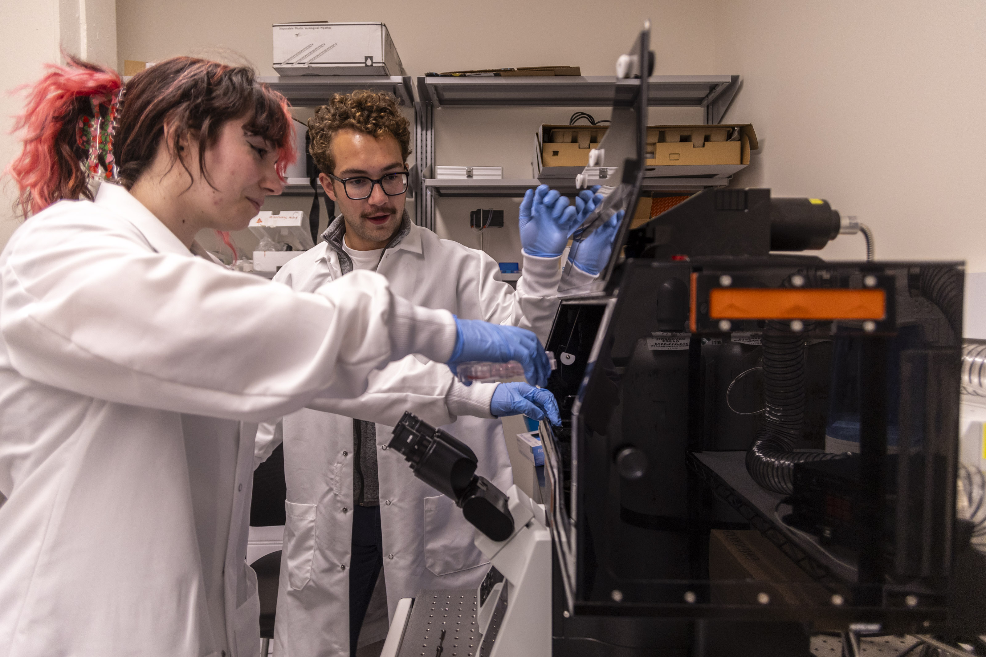 Two laboratory technicians in white coats and gloves working with scientific equipment.