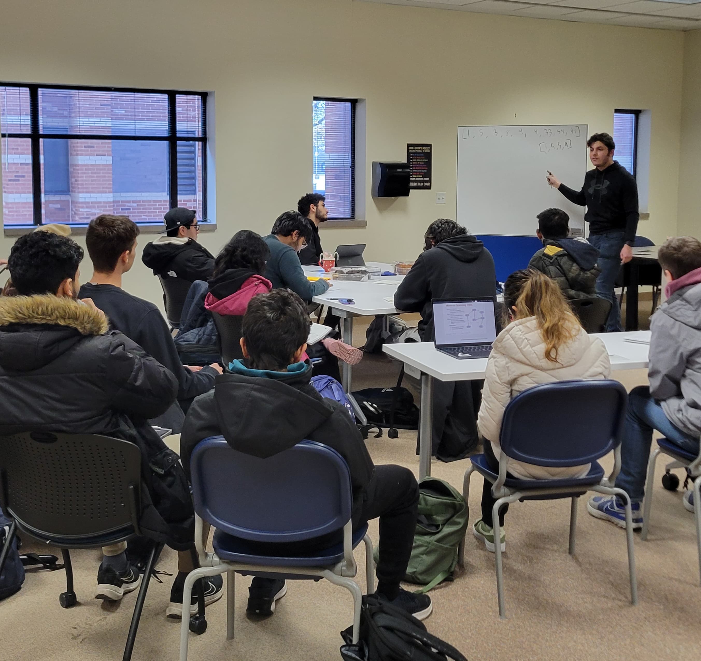 A classroom scene with a teacher standing near a whiteboard discussing with students seated at tables. Some students are using laptops. The room has a whiteboard with writings and windows in the background.