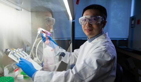 Wearing a white lab coat and protective equipment, Assistant Professor poses for a portrait while working in his cancer research lab