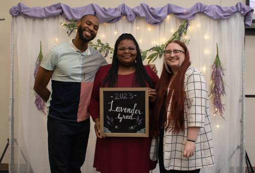 Three people standing in front of a white backdrop with a purple fabric top. They are holding a chalkboard sign that says "Lavender Grad"